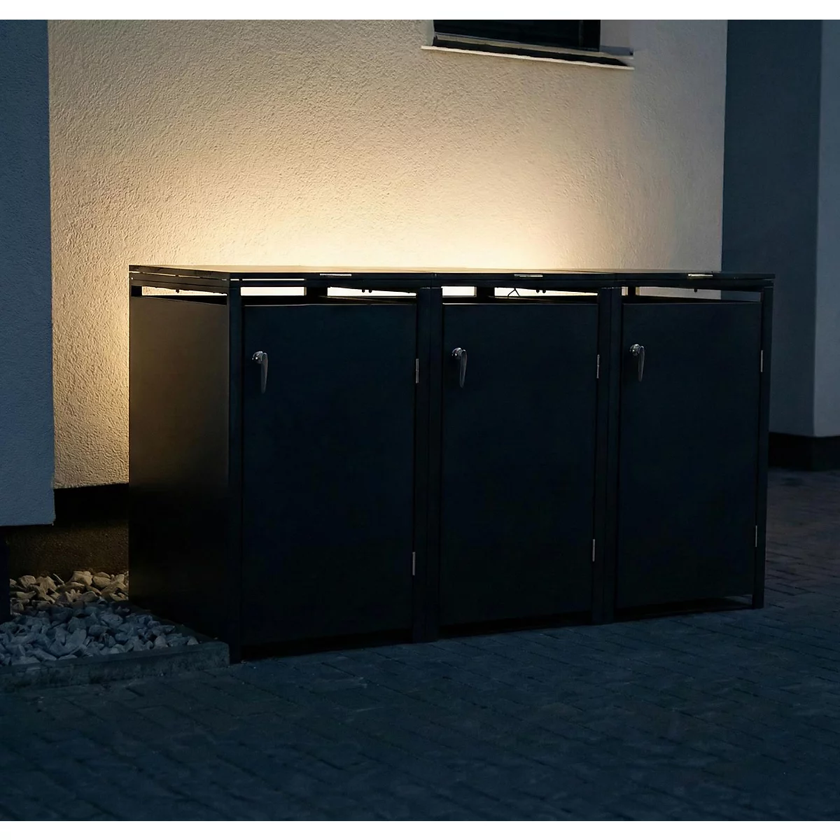 Donkere vuilnisbakkenkast met drie compartimenten voor een witte huismuur. Boven de bakken zijn lampen gemonteerd. Grind ligt ervoor.
