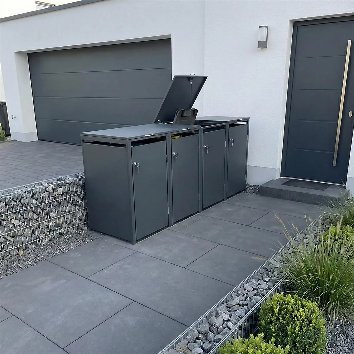 Donkergrijze vuilnisbakkenbox voor het huis. Eén bak is open, naast de voordeur en garage.