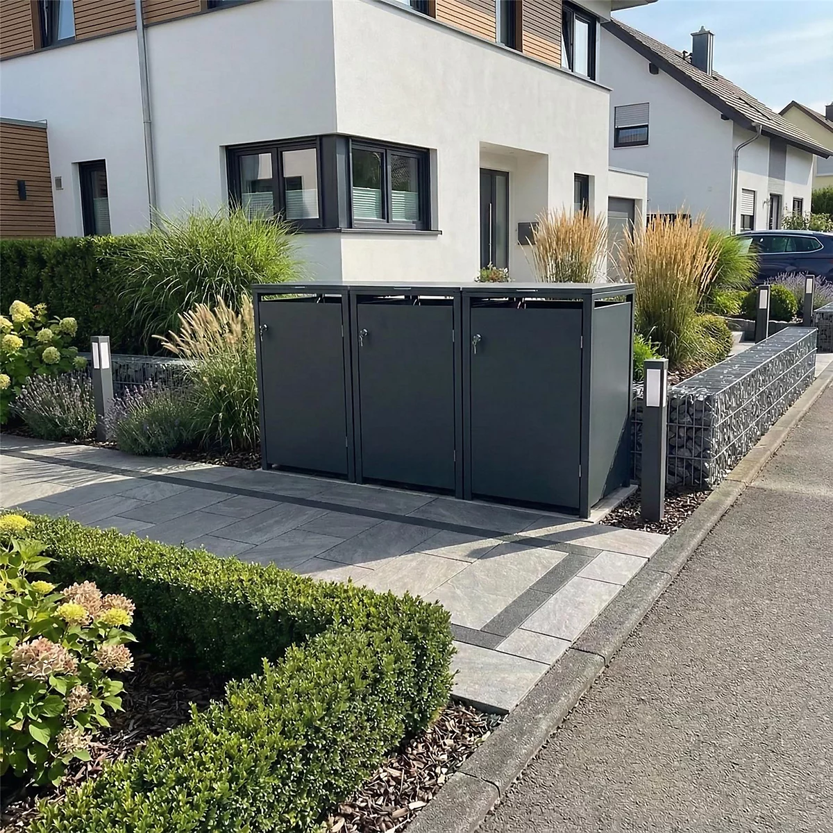 Drei graue Mülltonnenboxen vor einem Haus. Die Boxen sind von Grünpflanzen umgeben. Im Hintergrund ist ein Haus mit moderner Architektur zu sehen.