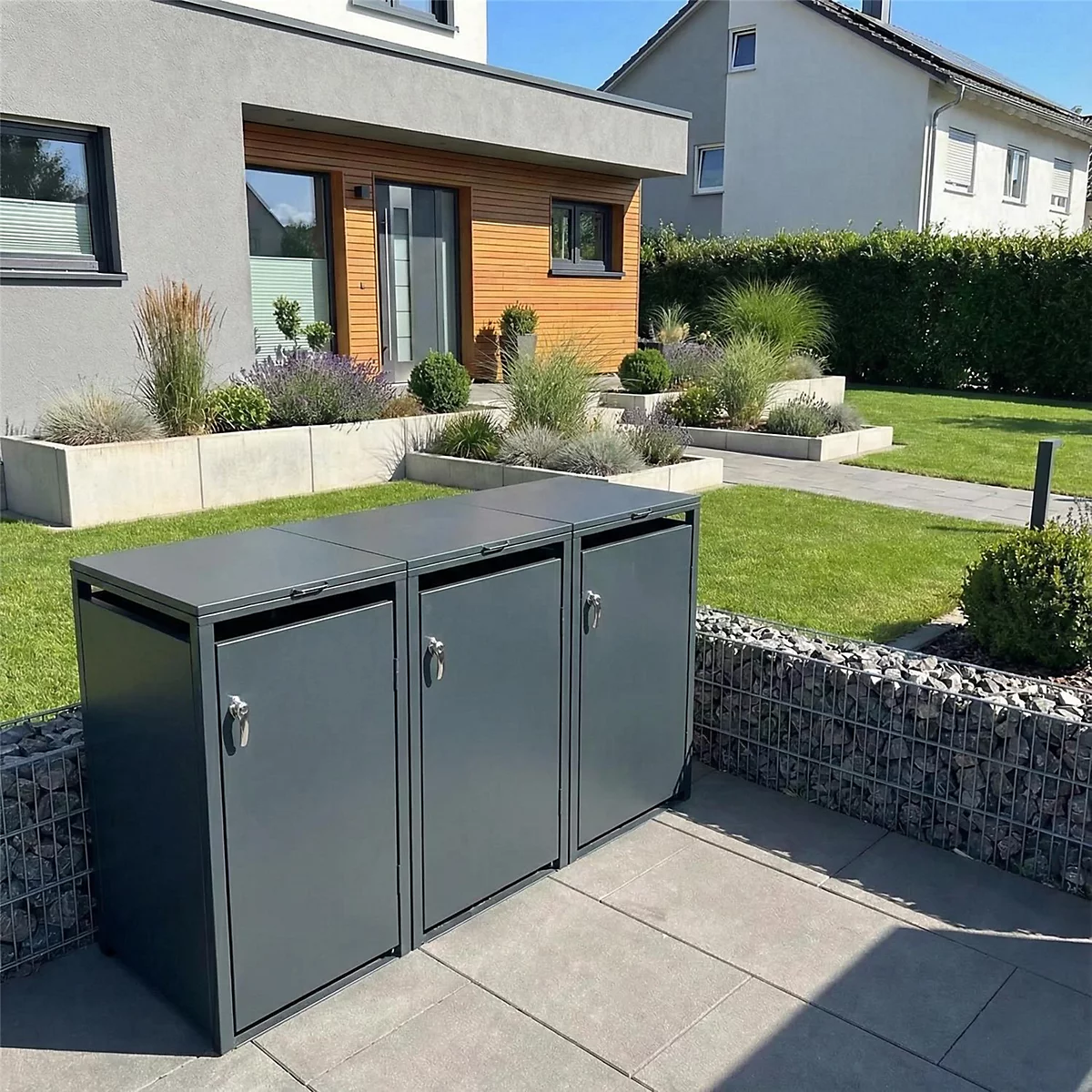 Drei graue Mülltonnenboxen auf einer Terrasse vor einem Haus. Im Hintergrund ist ein gepflegter Garten mit Haus zu sehen.