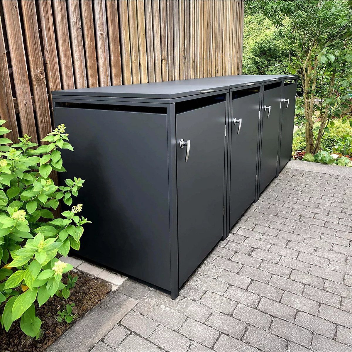 Drei graue Mülltonnenboxen im Garten, vor Holzwand und Sträuchern. Klinken und Pflastersteine.