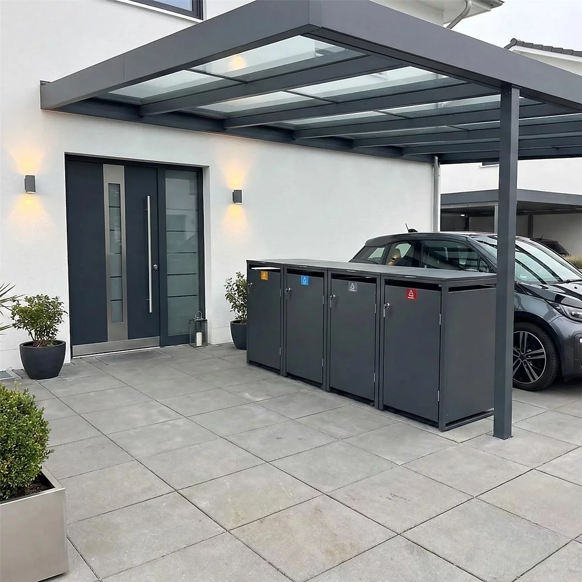 Dunkelgraue Mülltonnenbox unter einem Carport, vor einer Haustür. Das Bild ist aus der Perspektive des Betrachters aufgenommen.