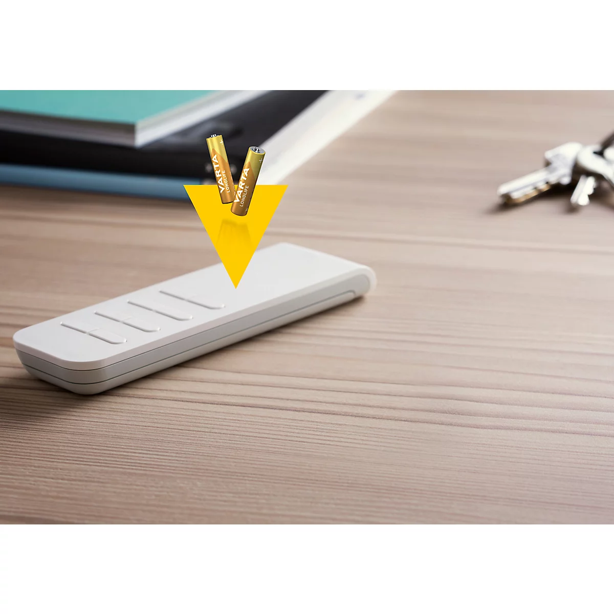 Batteries falling into a white device on a wooden table, keys and books in the background.