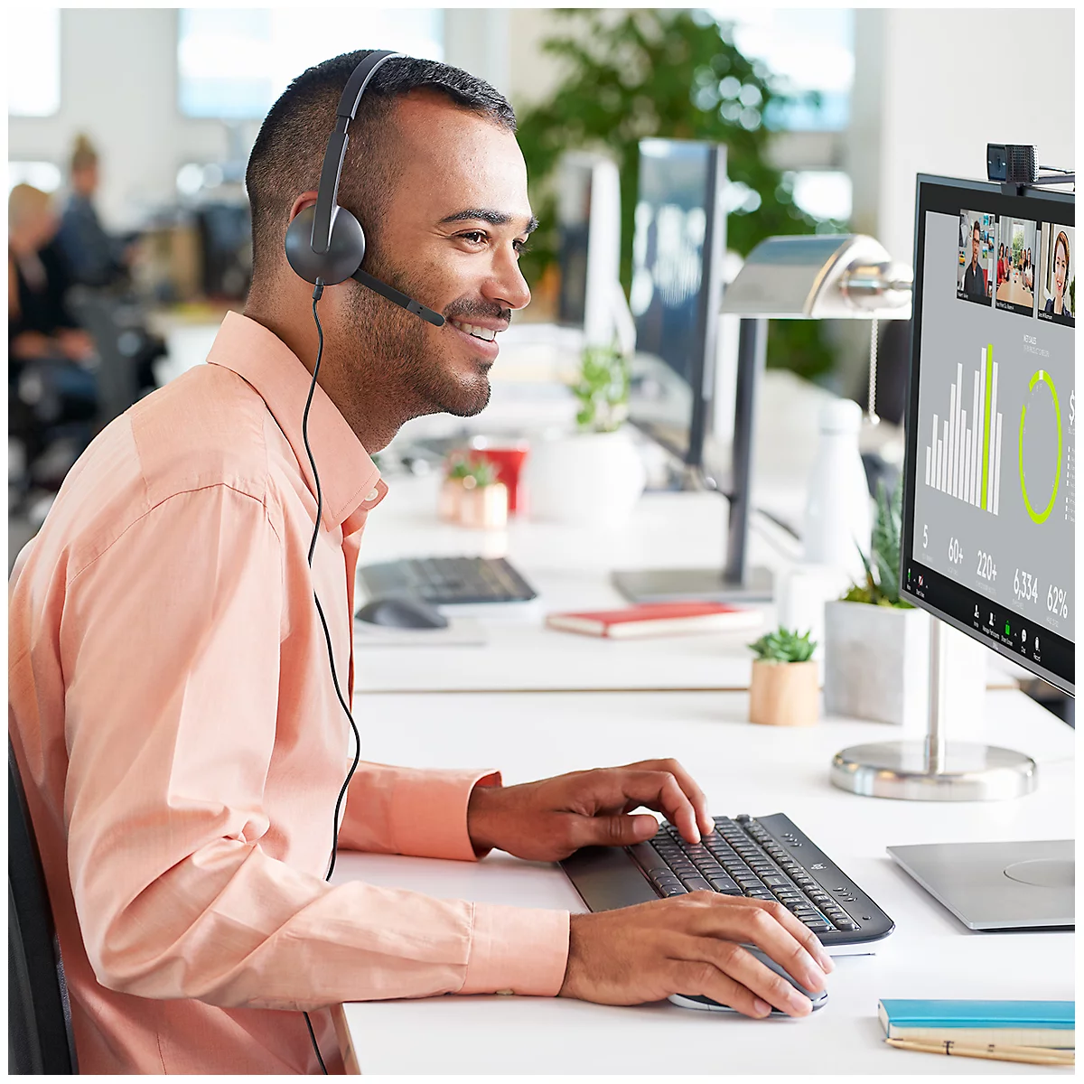 Un homme souriant avec un casque est assis à un bureau, regardant son ordinateur et travaillant. Un appel vidéo est visible à l'écran.