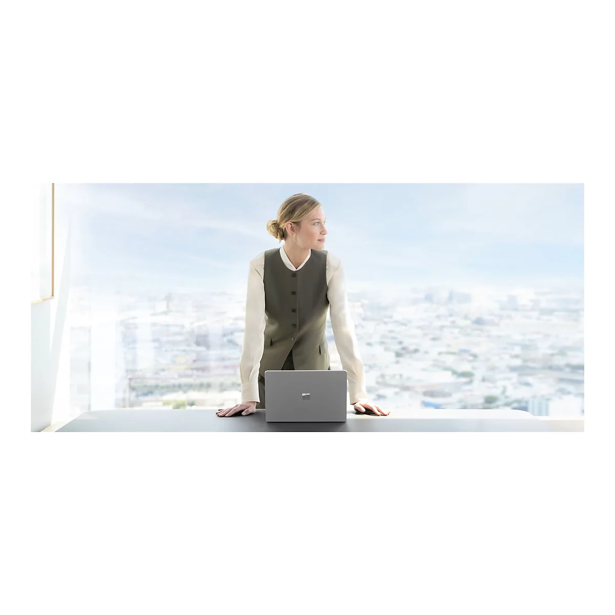 Eine Frau im BÃ¼ro, steht an einem Fenster und blickt auf die Stadt. Auf dem Tisch liegt ein Laptop.