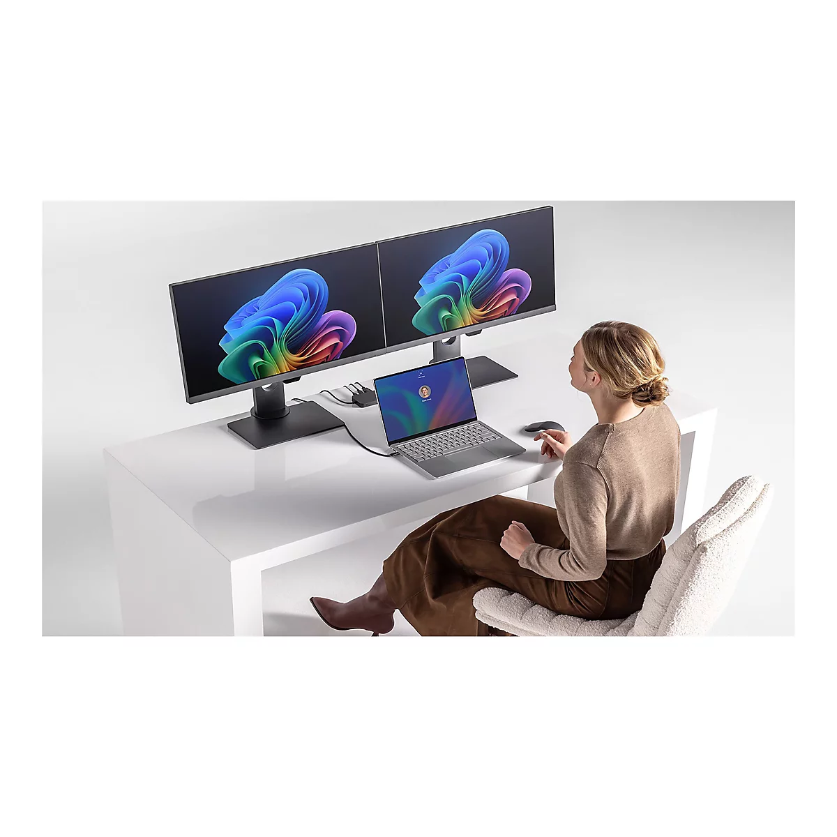 Eine Frau sitzt vor einem Schreibtisch mit zwei Monitoren und einem Laptop. Die Monitore und der Laptop zeigen bunte Grafiken.