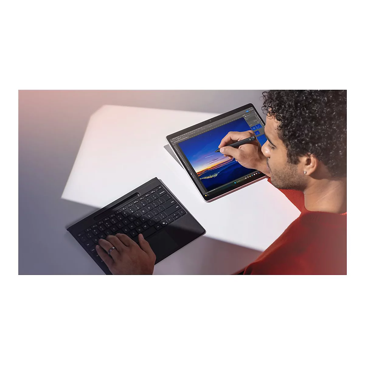 Mann zeichnet mit Stift auf Tablet. Auf dem Tisch liegt eine Tastatur.