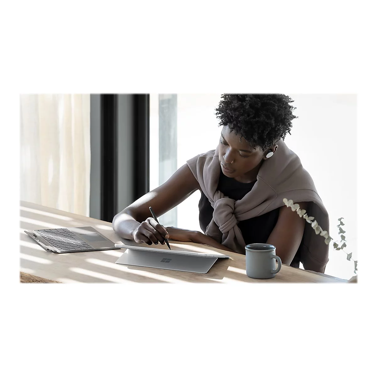 Une femme noire Ã©crit avec un stylo sur une tablette. Sur la table: tasse, ordinateur portable. ArriÃ¨re-plan: fenÃªtre, bois.