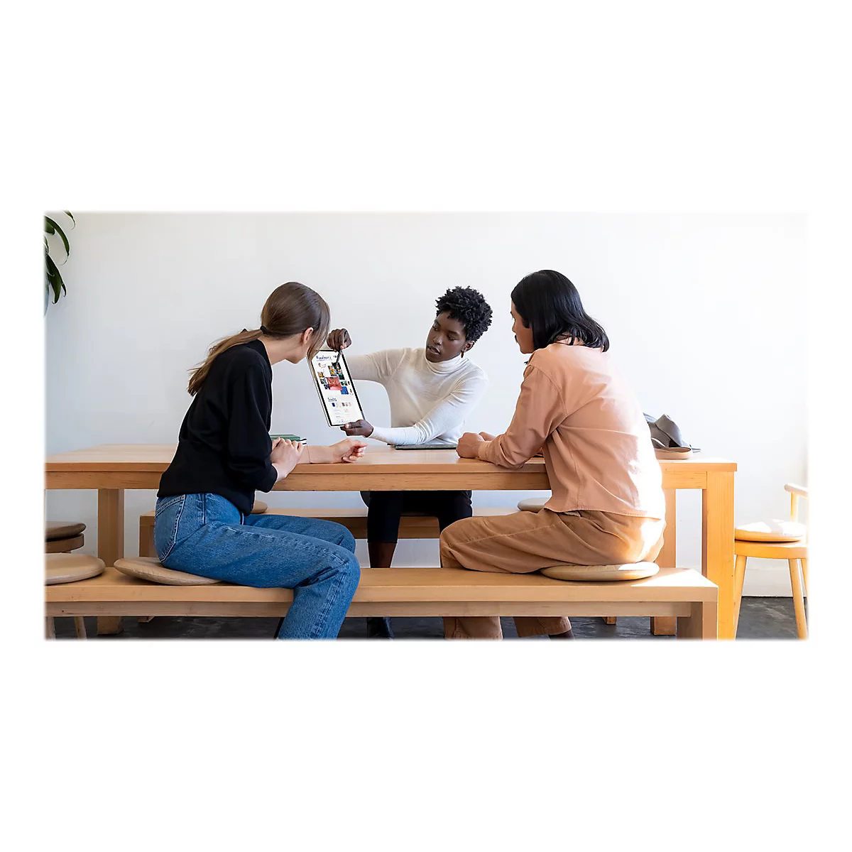 Trois femmes autour d'une table regardant une tablette. Une personne montre l'Ã©cran. Environnement lumineux.