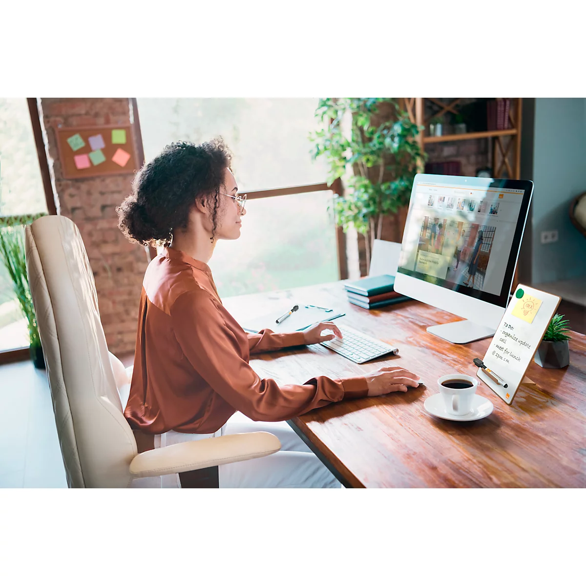 Une femme est assise à un bureau et travaille sur un ordinateur. Sur le bureau, il y a une tasse de café, un bloc-notes et un stylo.