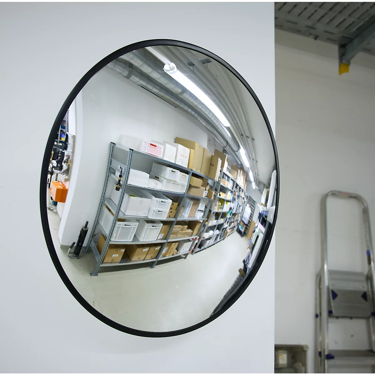 Miroir convexe rond avec cadre noir sur un mur blanc, reflétant des étagères d'entrepôt.