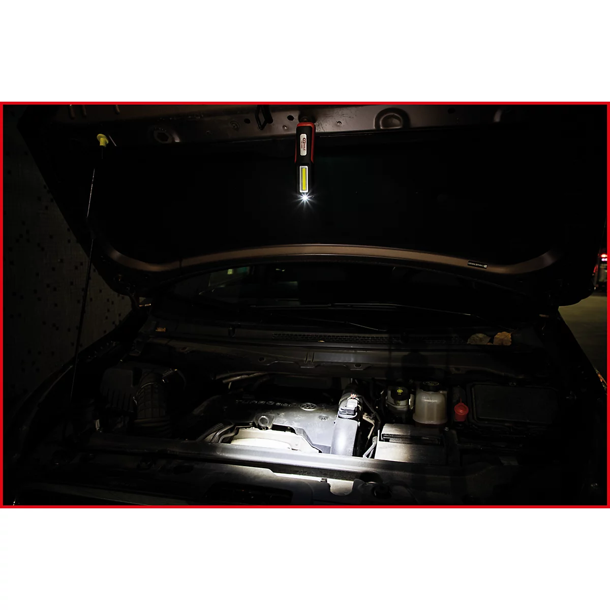 Ein offener Motorraum eines Autos wird von einer Stablampe ausgeleuchtet. Die Lampe hängt an der Motorhaube.