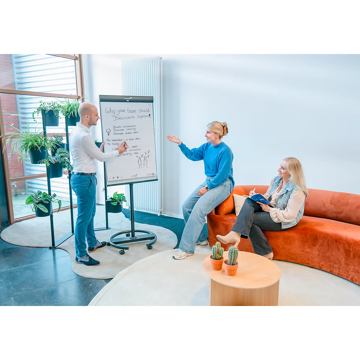 Drei Personen in einem Büro: Eine Person schreibt auf ein Whiteboard, zwei Frauen sitzen auf einem Sofa und diskutieren.