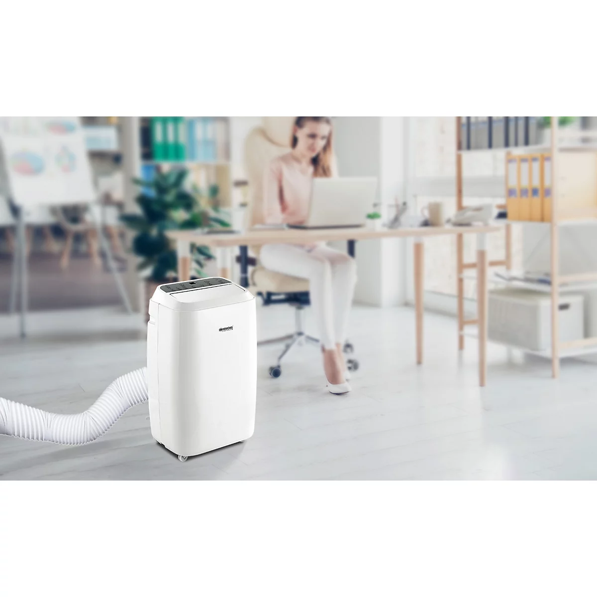 Ein weisser mobiler Klimaanlage steht im Büro. Im Hintergrund sitzt eine Frau an einem Schreibtisch.