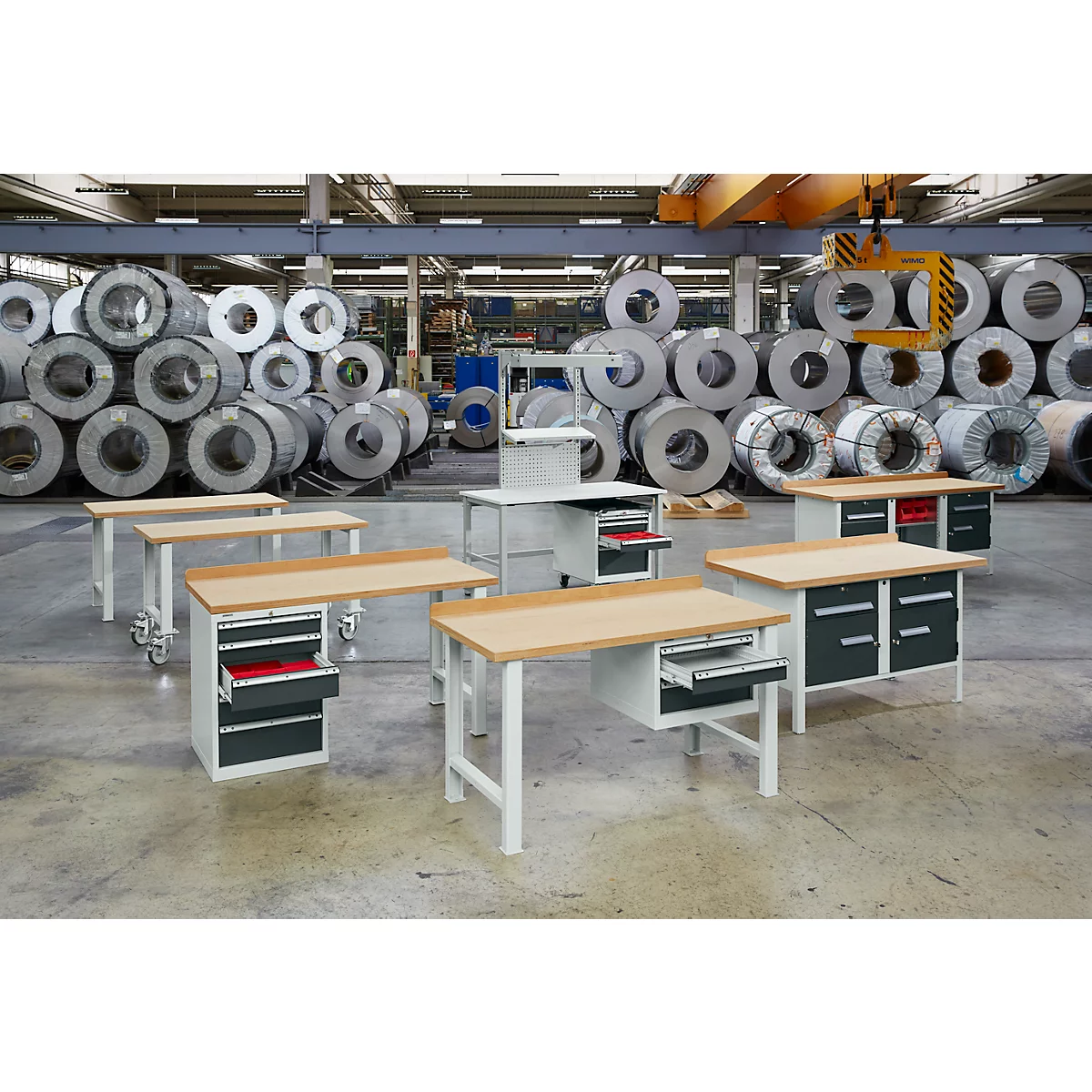 Workbenches in a factory hall. Behind the tables are large metal rolls stacked.