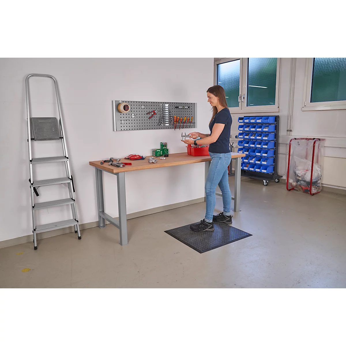 A woman stands at a workbench, working. Tools are mounted on a pegboard on the wall.