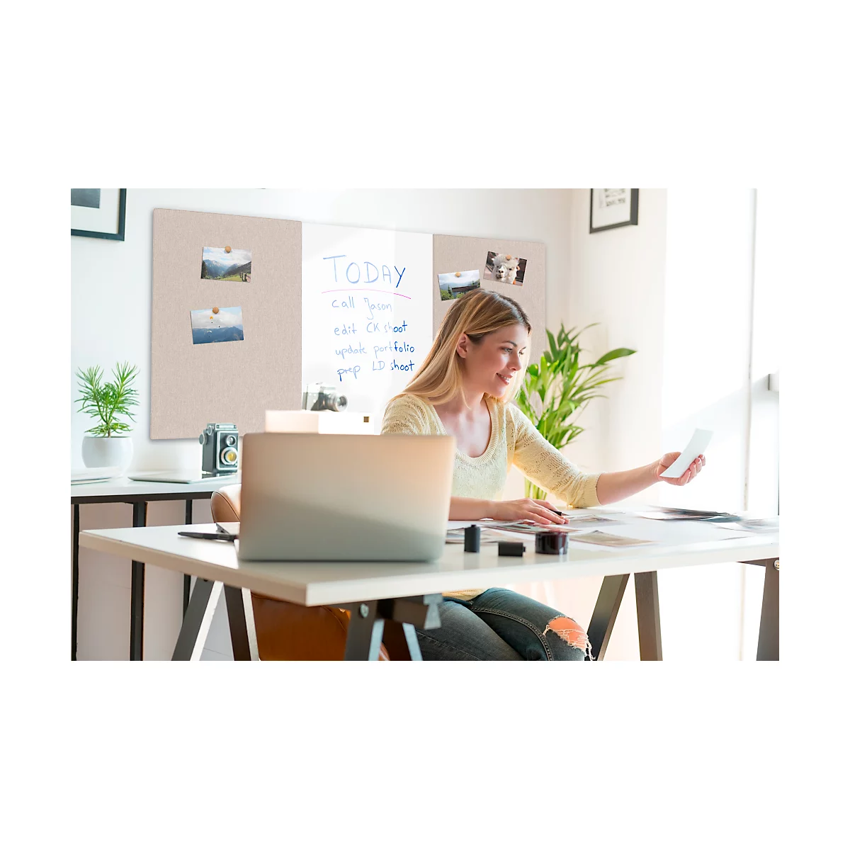 Une femme souriante est assise à un bureau avec un ordinateur portable, tenant et regardant une photo. En arrière-plan, un tableau d'affichage et des plantes.