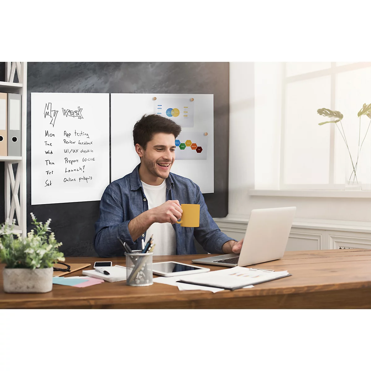 Un homme est assis à un bureau, boit du café et travaille sur un ordinateur portable. Sur un tableau, il y a des notes de travail.
