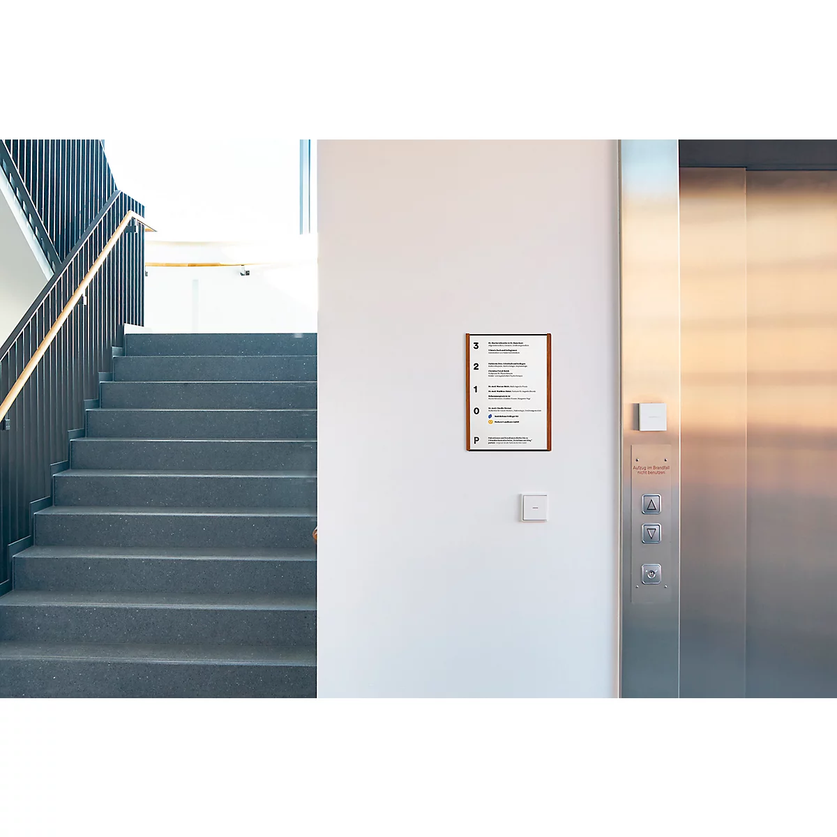 Escalier gris, support marron, panneaux avec inscriptions sur mur blanc. Ascenseur à droite.