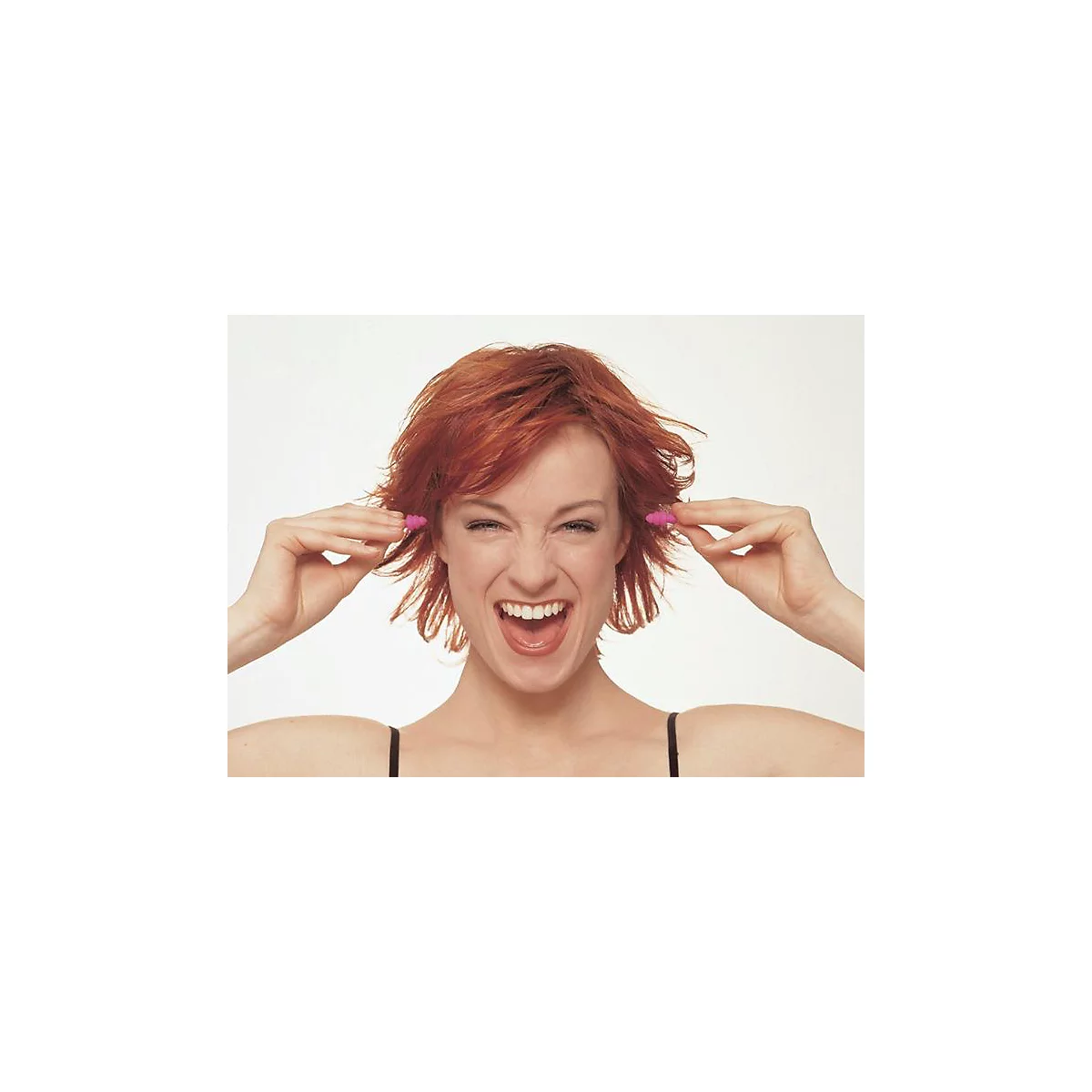 Une femme aux cheveux roux et aux bouchons d'oreille roses sourit en se tenant les oreilles.