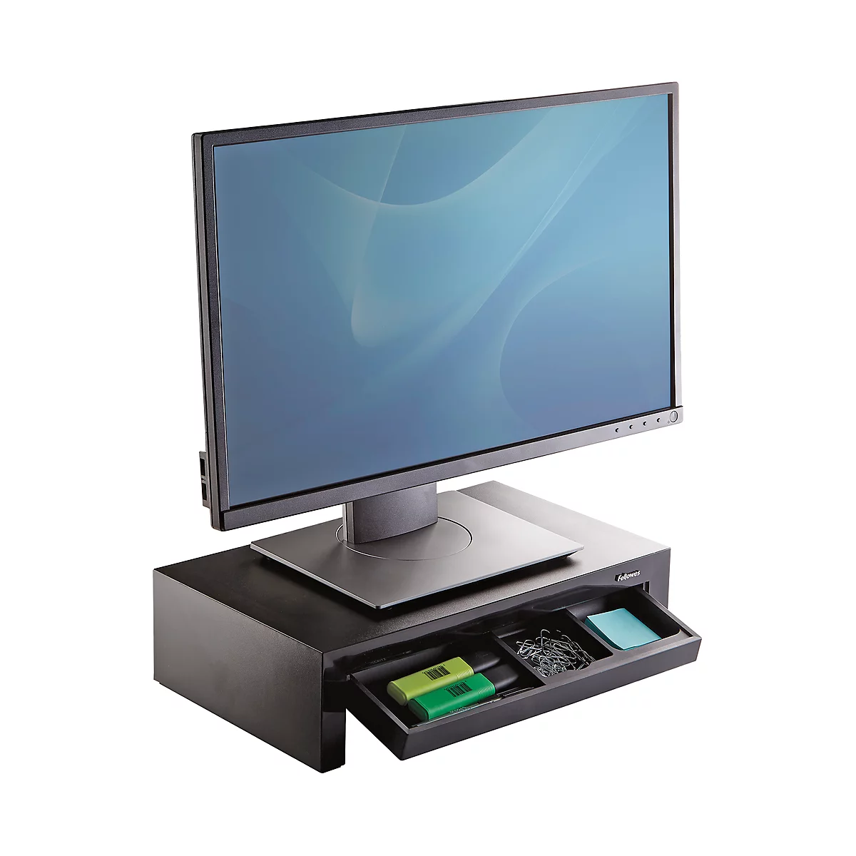 A black monitor stand with a drawer, on which a screen stands, there are pens and paper clips in the drawer.