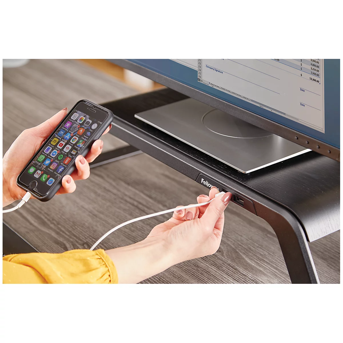 Person connects smartphone with charging cable to a monitor stand. Screen with applications, yellow top.
