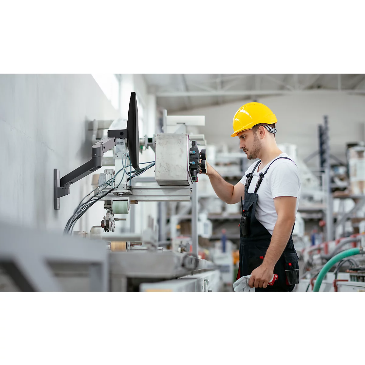 Ein Mann mit gelbem Helm arbeitet an einer Maschine in einer Fabrik. Er trägt eine Latzhose und ein weißes T-Shirt.