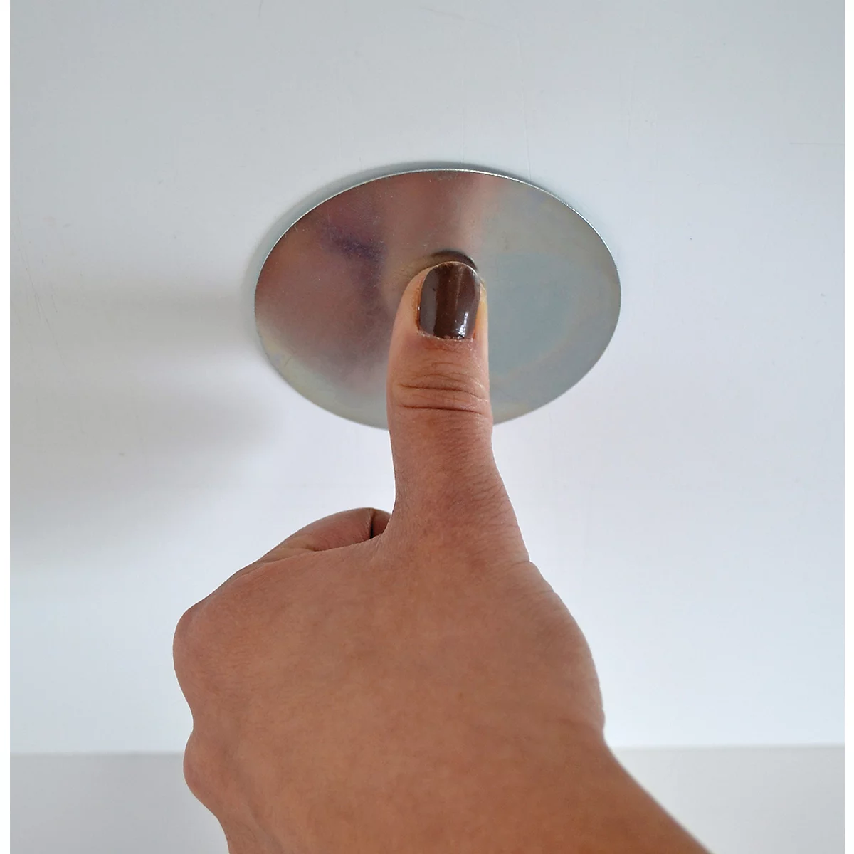 Eine Hand mit braun lackiertem Fingernagel drückt auf eine metallische Scheibe an der Decke.