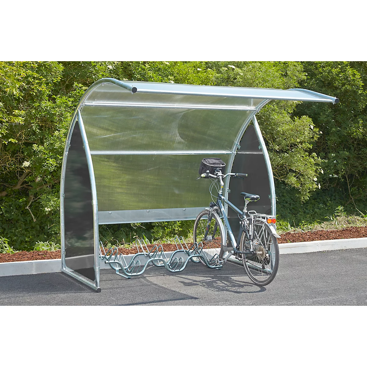 Bicycle stands under a transparent shelter with a metal frame. Trees in the background.