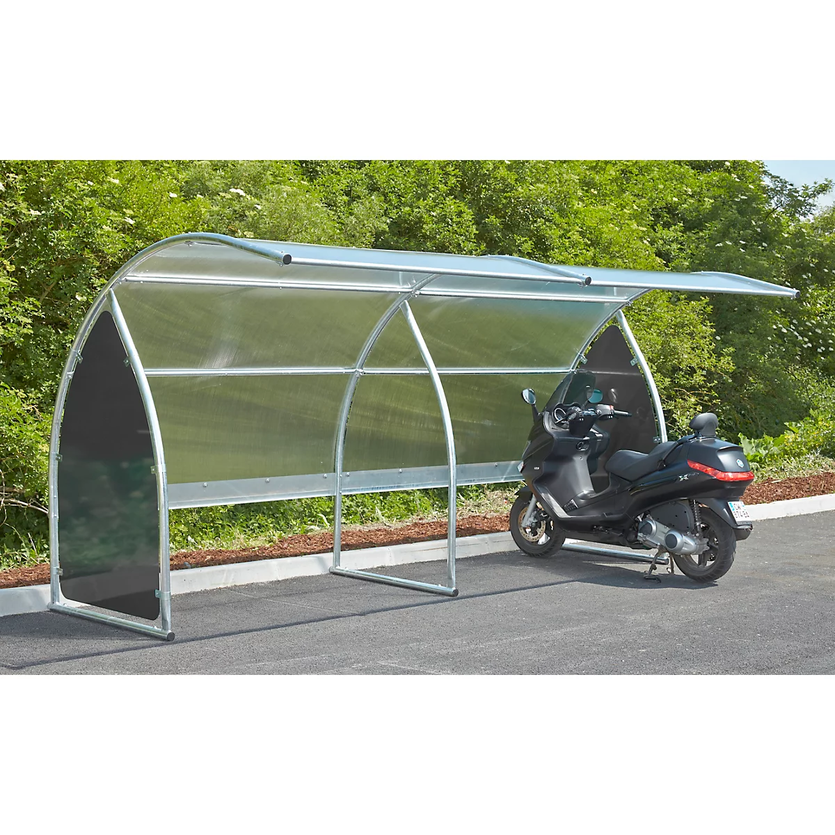 Motorcycle under a metal shelter on asphalt. Green bushes in the background.