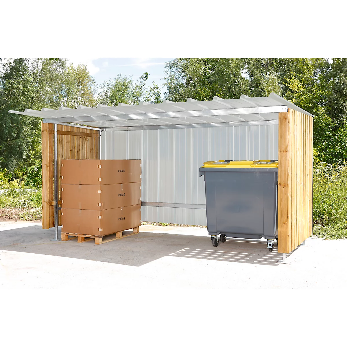 A shelter with a trash can and cardboard boxes. Gray bin with yellow lid, cardboard boxes on a pallet.