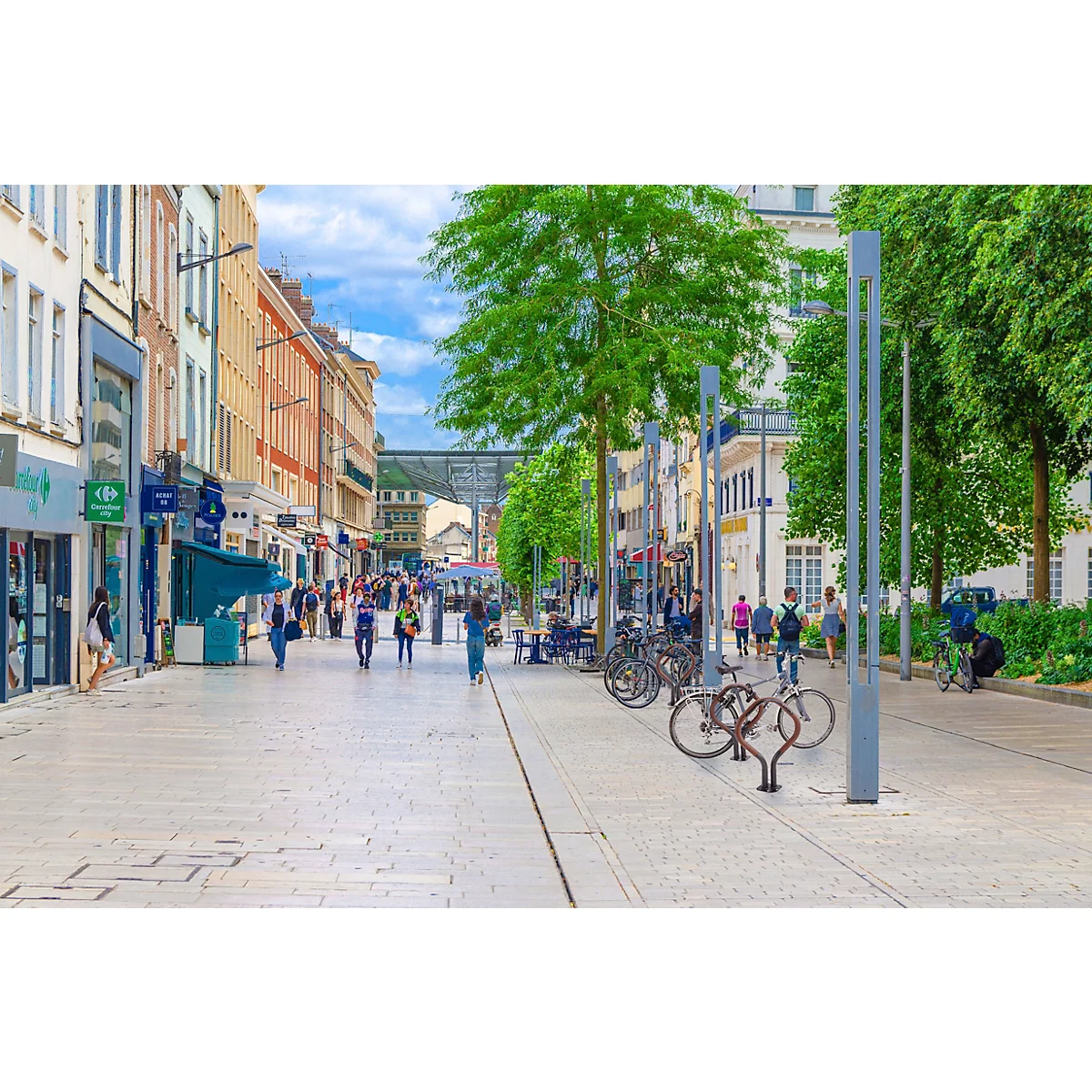 A busy shopping street with shops, people and bicycles. Tall buildings line the paved street, trees provide shade.