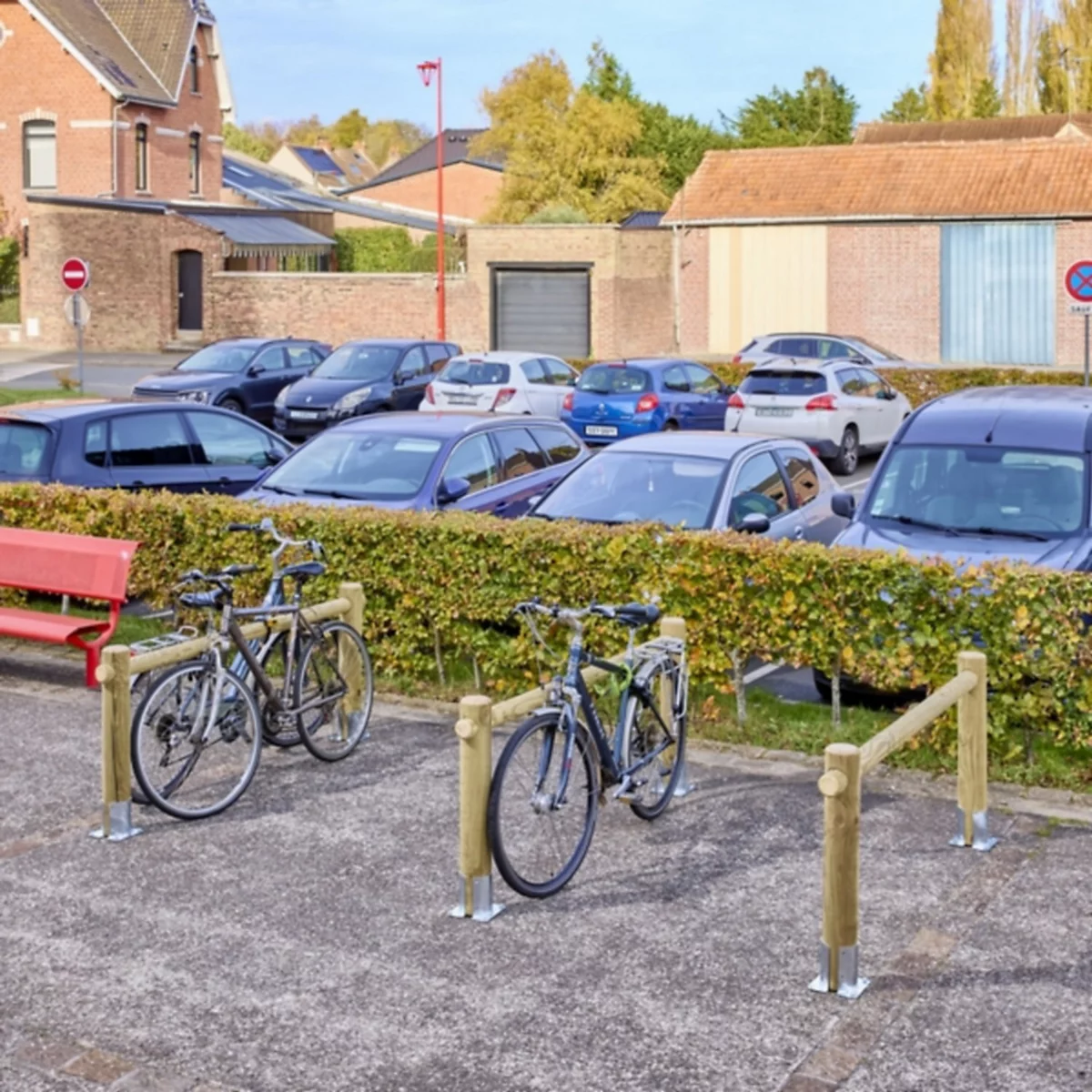 Zwei Fahrräder stehen in einem hölzernen Fahrradständer auf einem gepflasterten Platz vor einer Hecke und einem Parkplatz.