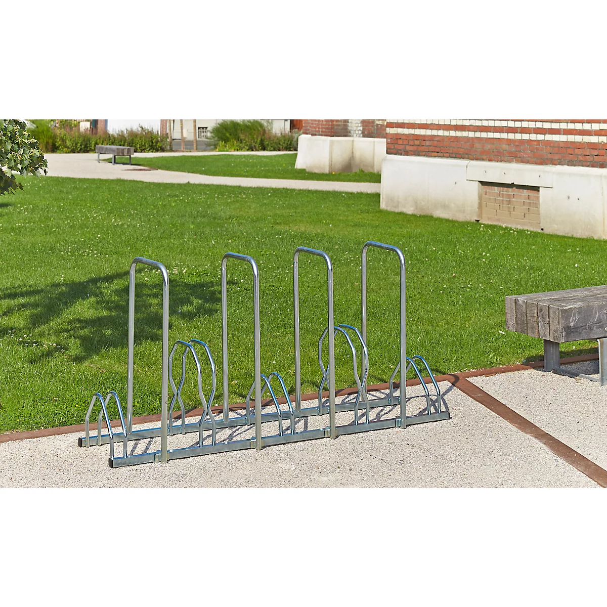 Metalen fietsenrek op verharde ondergrond, daarachter gazon, gebouw.