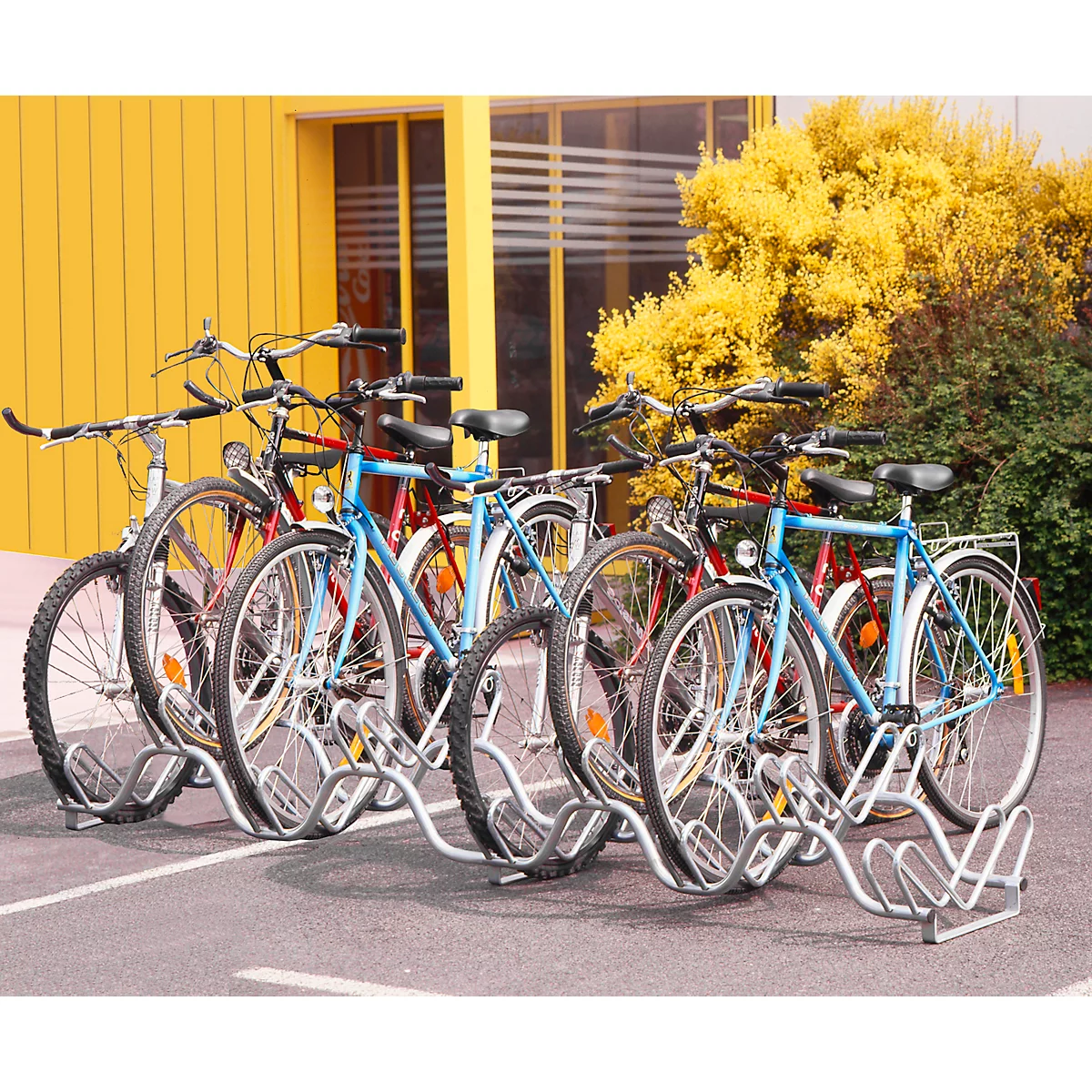 Verschillende fietsen geparkeerd in een buitenfietsenrek. Op de achtergrond een geel gebouw en groene gebieden.