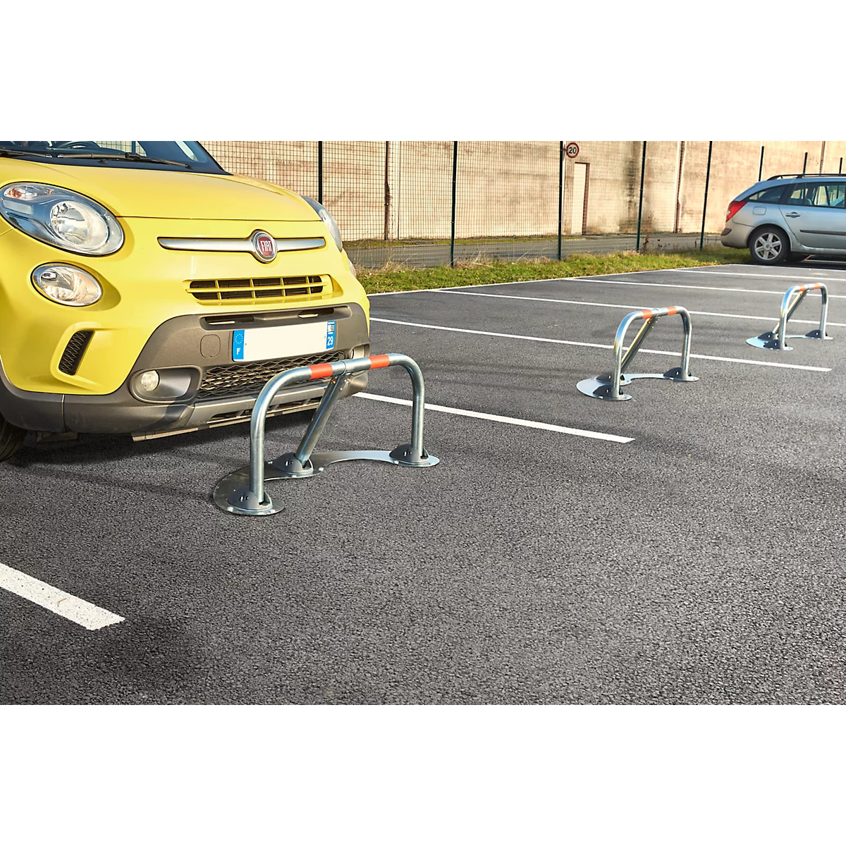 Yellow car parking behind parking barrier. In front, a second barrier, behind which parking bays are marked. Grey asphalt.