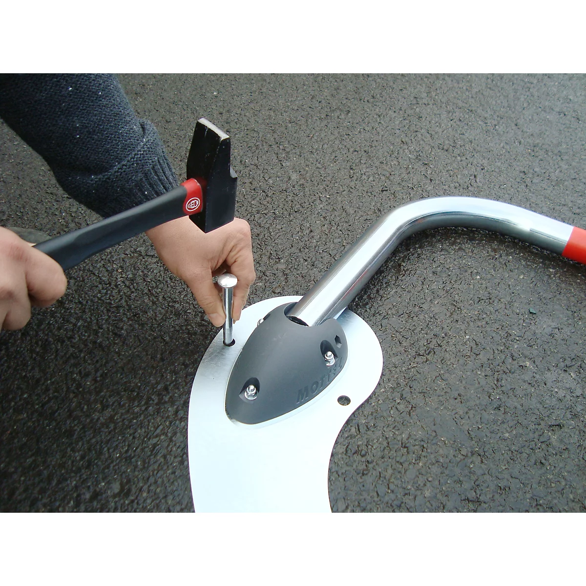 Person hammers a nail into a metal plate. Next to it is a curved metal tube with a red tip.