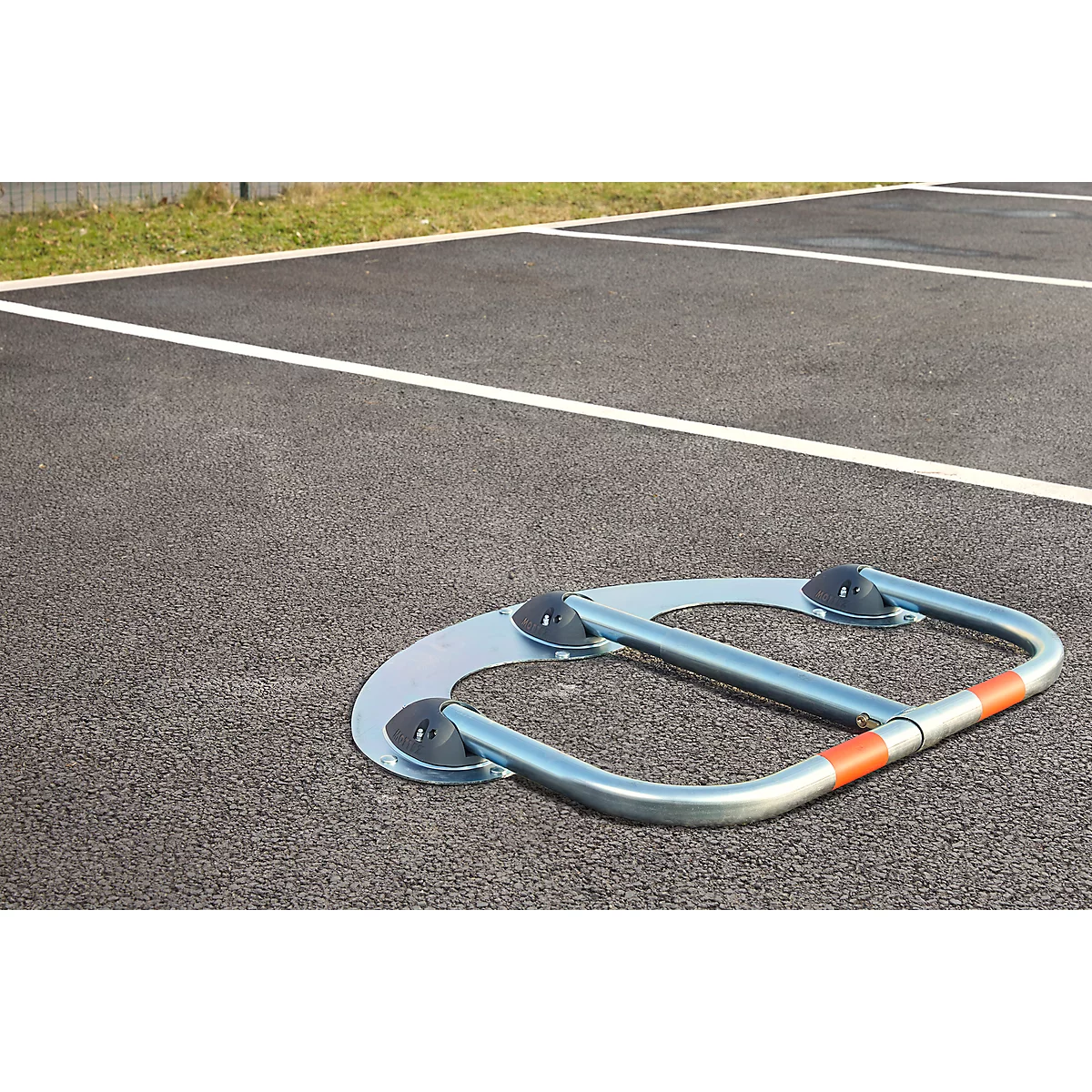 Metal parking barrier on asphalt. Silver, with orange stripes and black rubber feet attached.