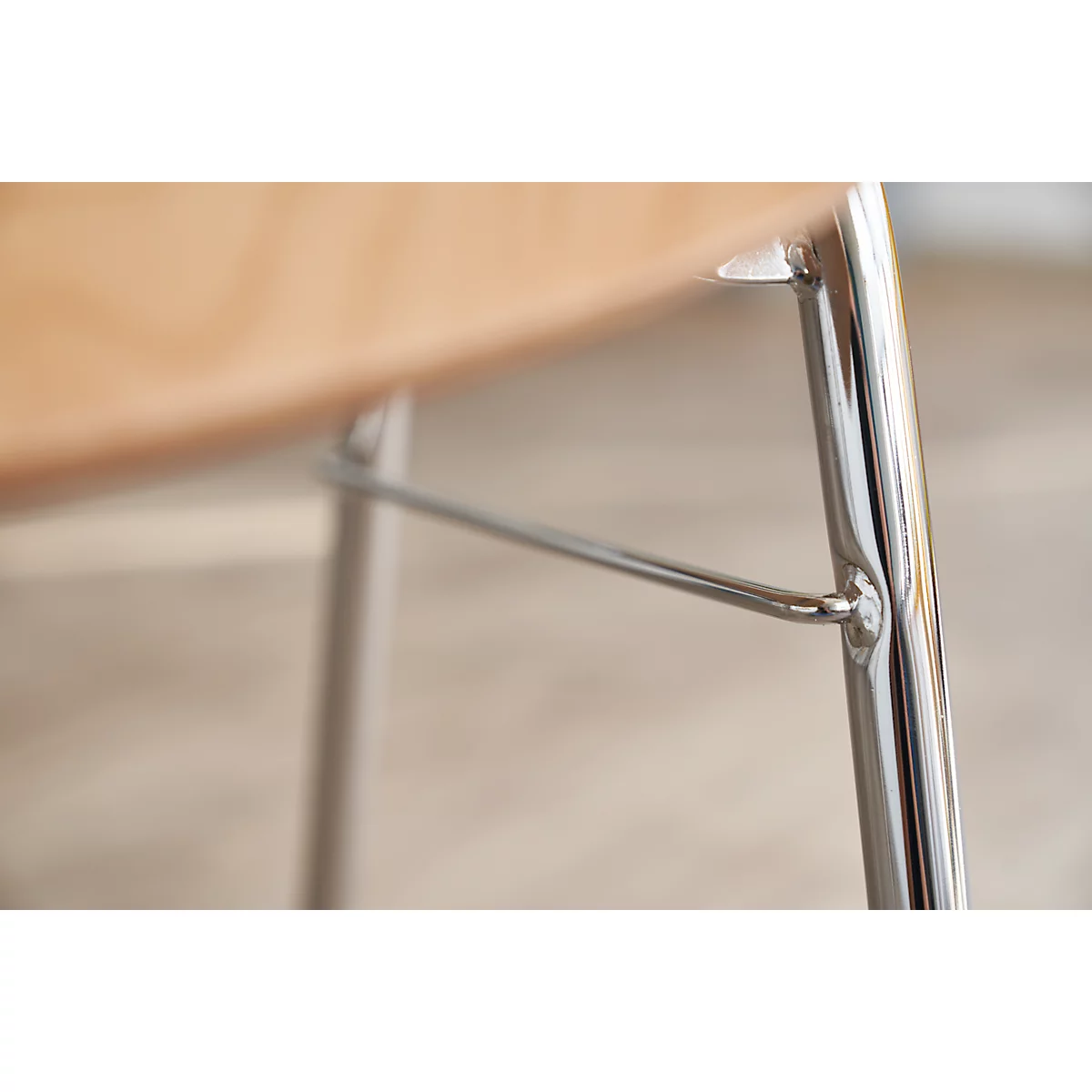 Close-up of a chair leg. The legs are made of chrome-plated metal, connected with a horizontal strut. The seat is light brown, made of wood.