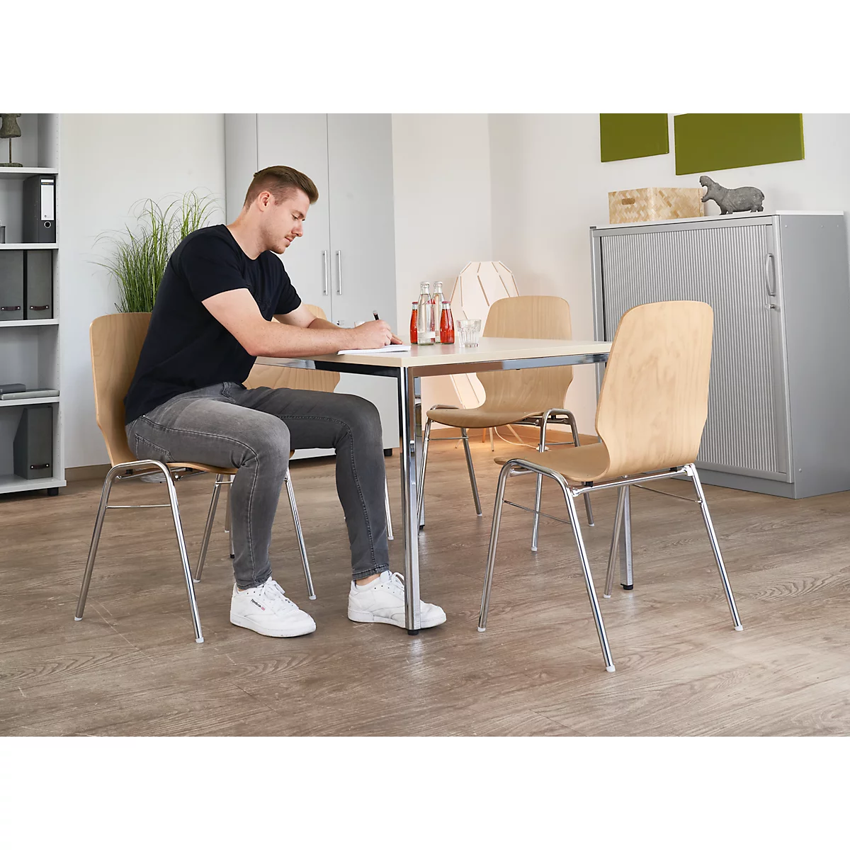 Man sitting at table, writing. Wooden chairs with metal legs. Light wood, grey trousers, black t-shirt, white sneakers.
