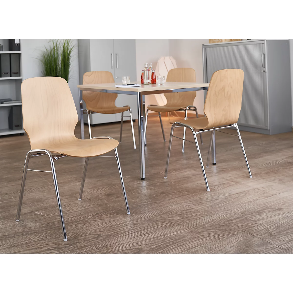 Four chairs with wooden backs and chrome legs stand around a table with drinks.