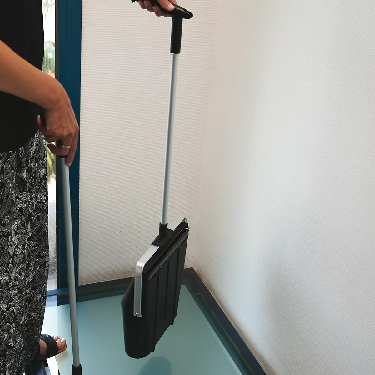 Person hält schwarze Kehrschaufel mit langem Stiel. Der Stiel wird mit einer Hand gehalten. Am Boden ist eine Glasplatte zu sehen.