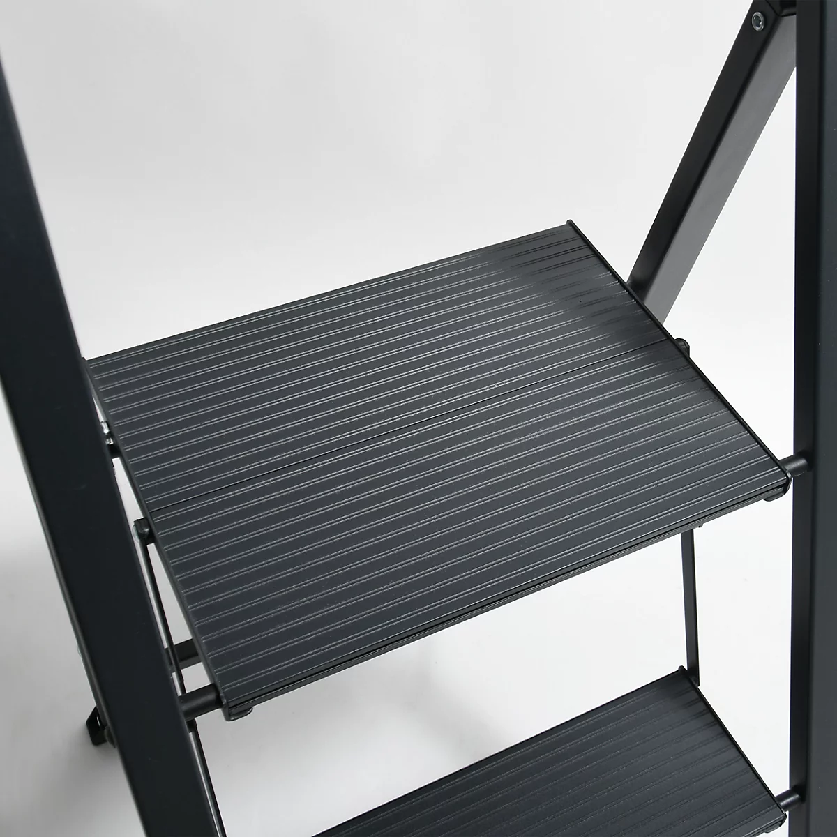 Detailaufnahme einer schwarzen Trittleiter. Die Stufen sind geriffelt, der Hintergrund ist hell.