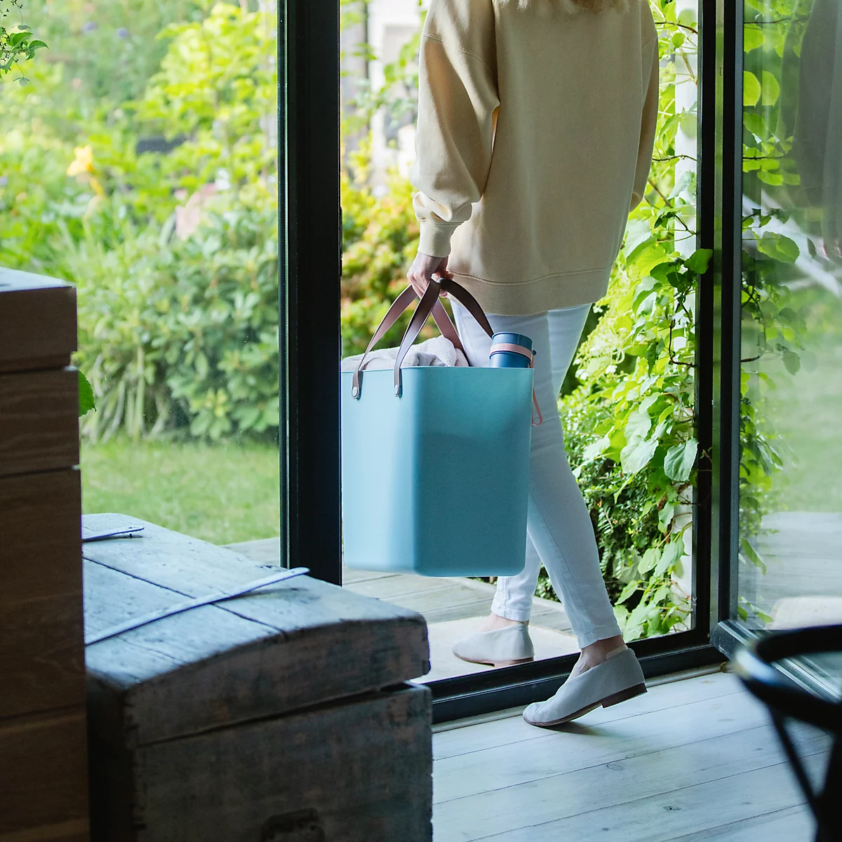 Person with light blue container steps into a garden. The container is filled with items. The person is wearing light clothing.