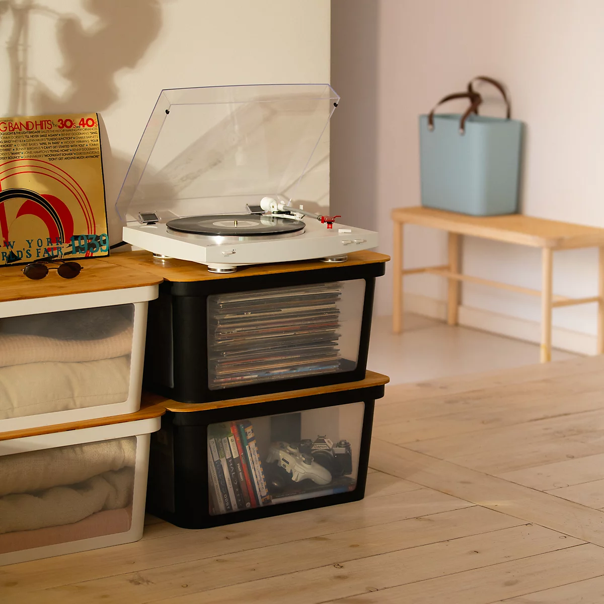 A record player sits atop a stack of clear boxes filled with records and books. Beside, a light bench and a blue bag.
