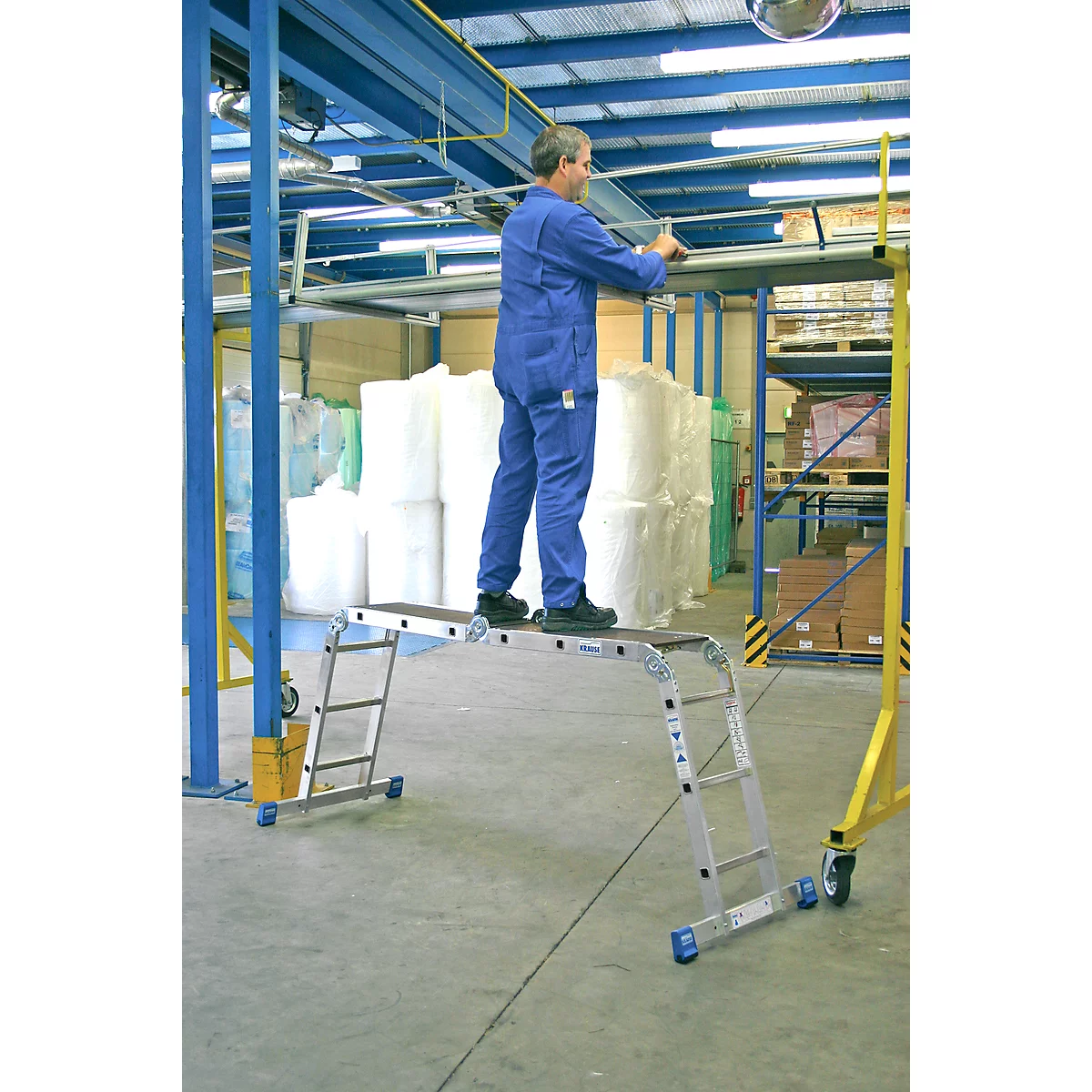 Man stands on a platform ladder and works in a factory. Materials and machines can be seen in the background.