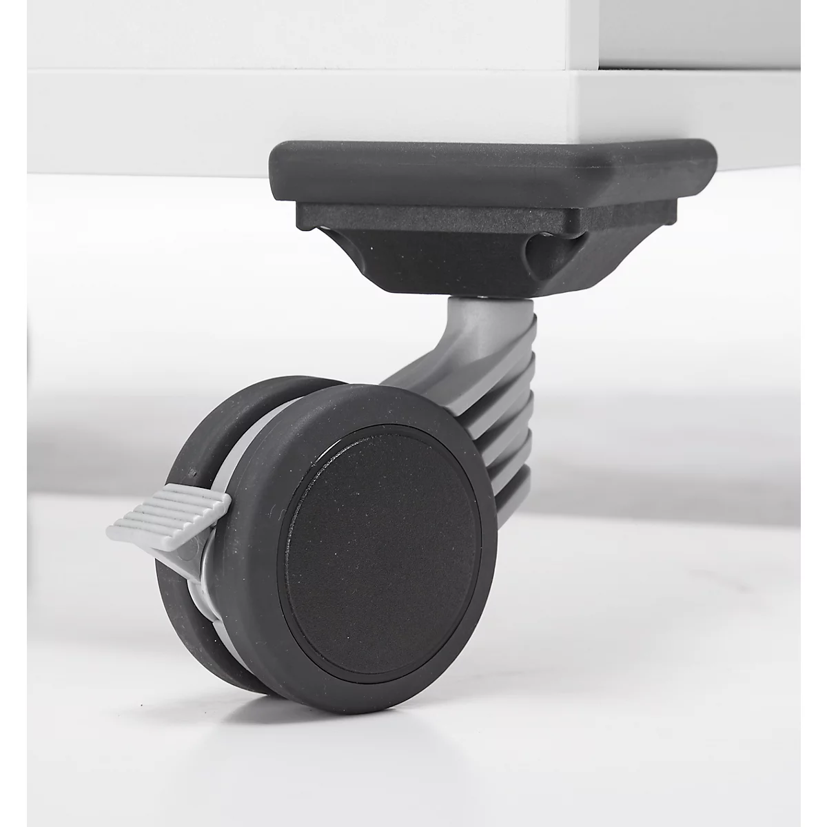 Close-up van een wiel met een zwart wiel en remhendel, onder een wit meubelstuk.