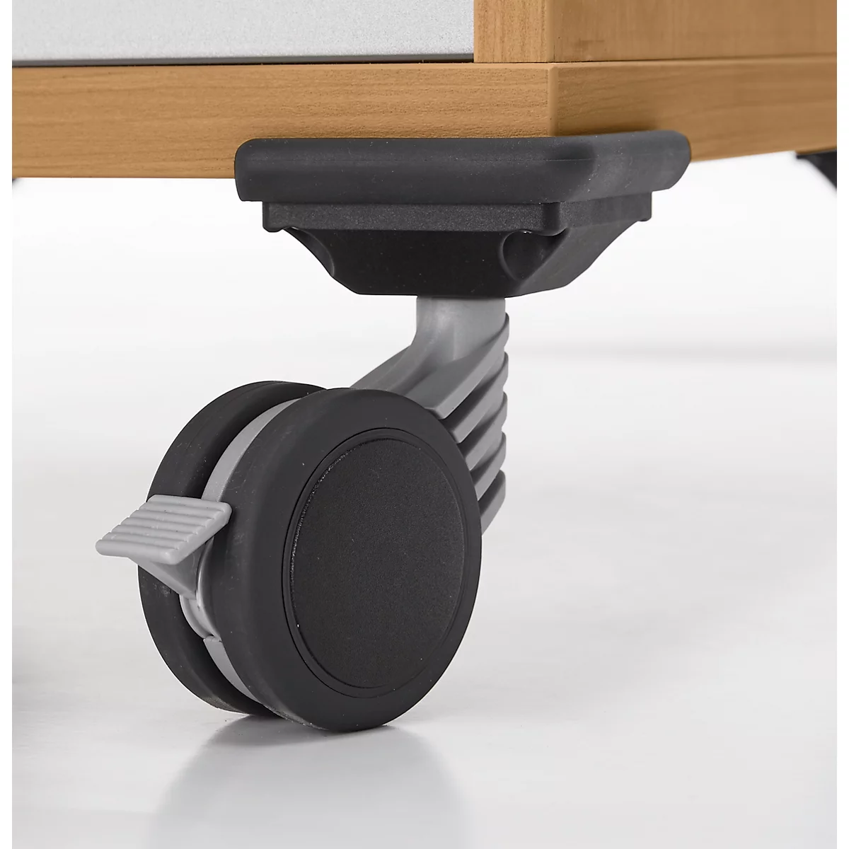 Nahaufnahme eines Möbelrads mit Feststellbremse. Das Rad ist schwarz, der Halter grau. Holzmöbel im Hintergrund.