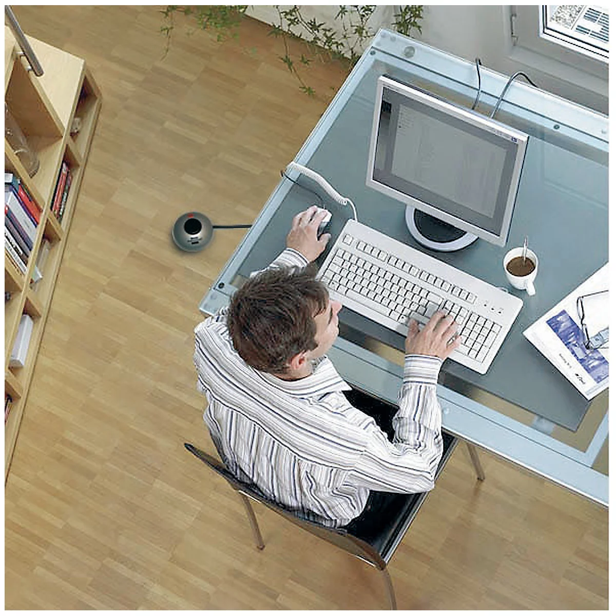 Un homme en chemise rayée est assis à un bureau en verre, travaillant sur un ordinateur. Une tasse de café est devant lui.