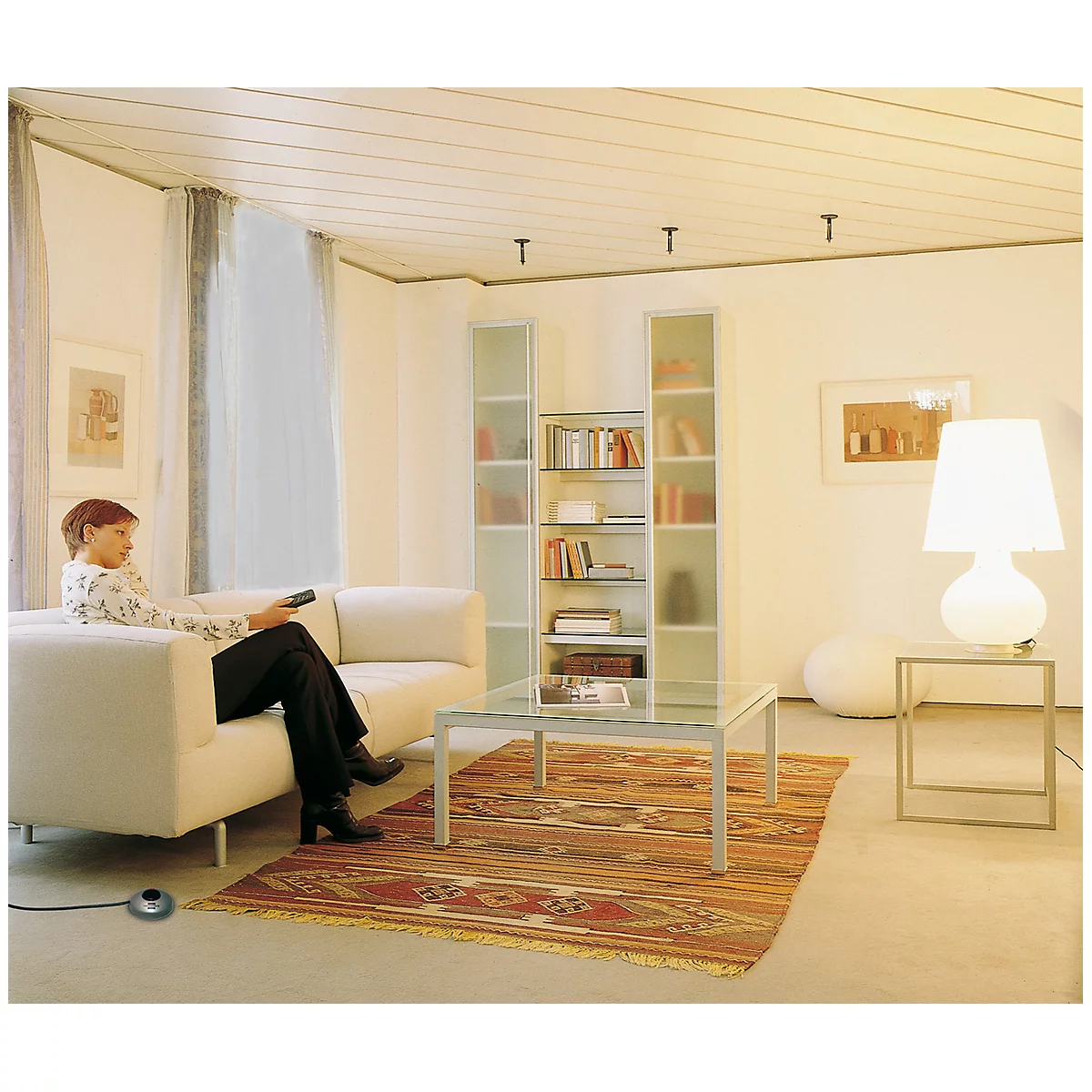 Une femme est assise sur un canapé blanc et regarde la télévision. La pièce est lumineuse avec un tapis beige et un grand abat-jour blanc à côté d'une table en verre.